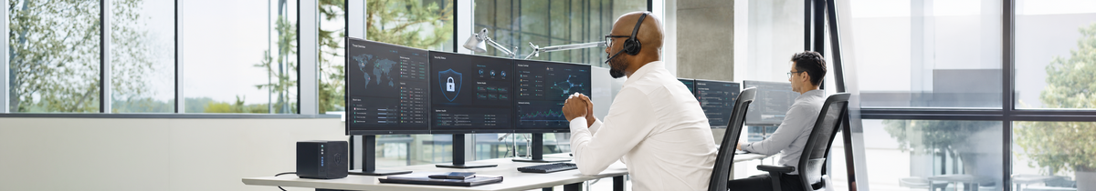 A man wearing a headset working at three LG monitors displaying security and network dashboards alongside an LG Thin Client device, with a colleague working in the background; Thin Clients tab selected in the product menu below