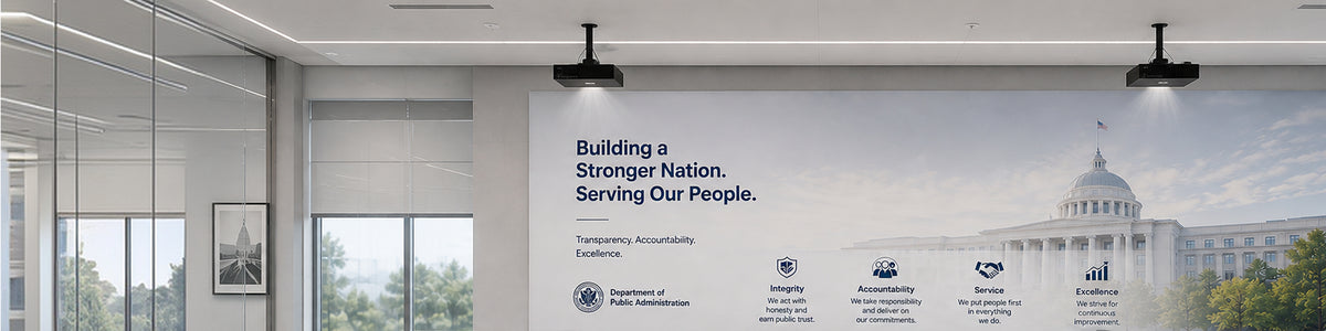 Two ceiling-mounted projectors display a government presentation on a large wall screen above a long desk with four monitors and office chairs.