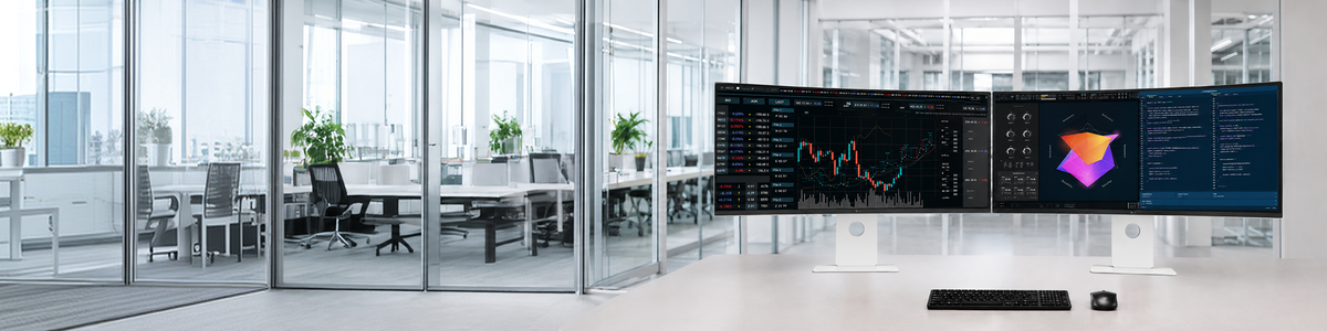 Two ultrawide monitors displaying various data graphs and code are placed side-by-side on a desk in a glass-walled office, with a black keyboard and mouse in front of them.