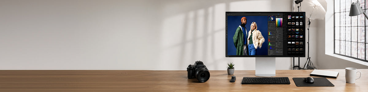 A monitor displaying photo editing software sits on a wooden desk alongside a camera, a small potted plant, a keyboard, and a mouse.